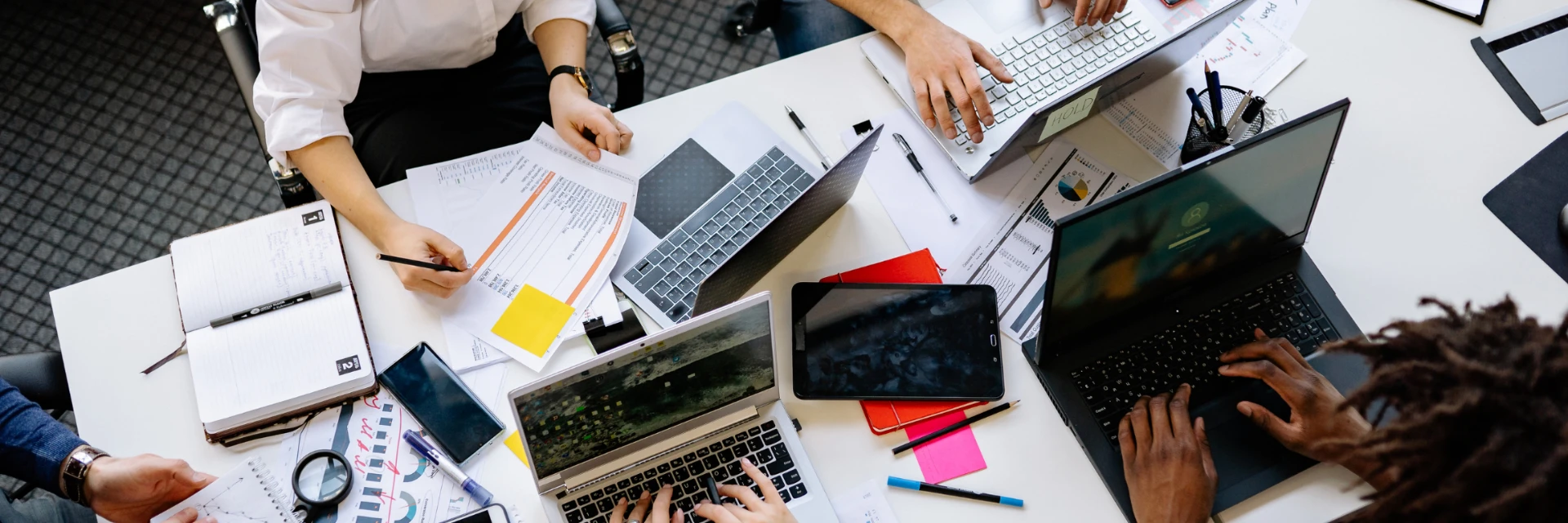 Ã‰quipe projet en vue plongeante collaborant autour d'une table encombrÃ©e d'Ã©crans et de documents.