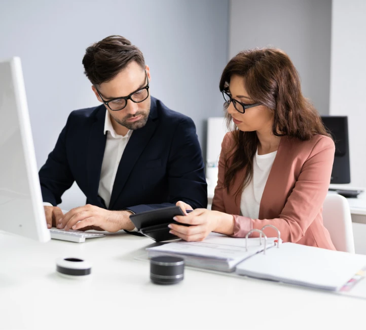 Un homme et une femme à lunettes travaillent ensemble sur un dossier et une calculatrice.
