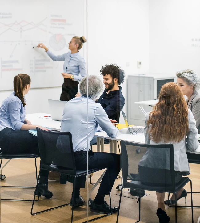 Réunion d'équipe : une femme présente des graphiques au tableau devant six collègues attentifs.