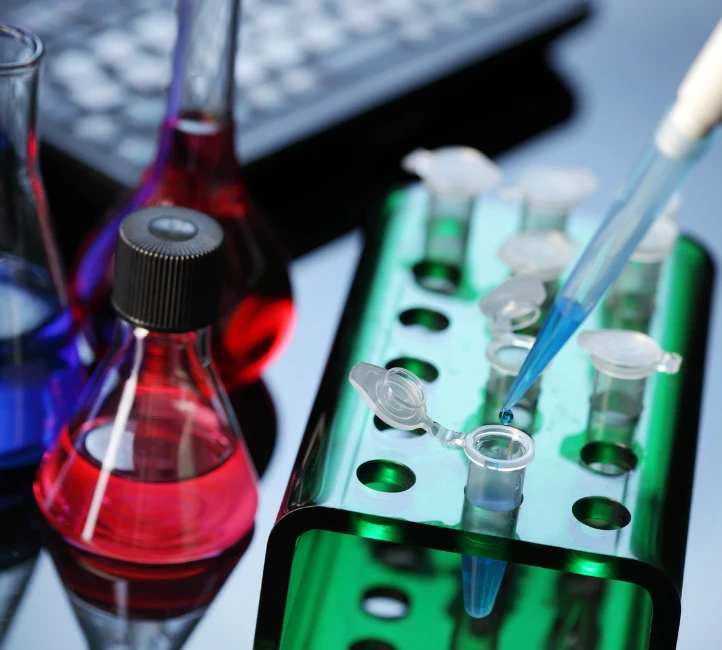 Pipette versant un liquide bleu dans un microtube, entourée de flacons de laboratoire colorés.