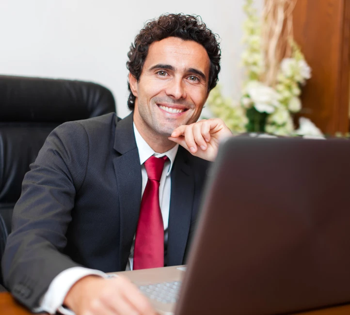 Homme souriant en costume et cravate rouge devant un ordinateur portable au bureau.