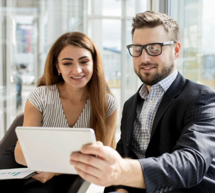 Un homme et une femme souriants regardent ensemble une tablette dans un bureau lumineux.